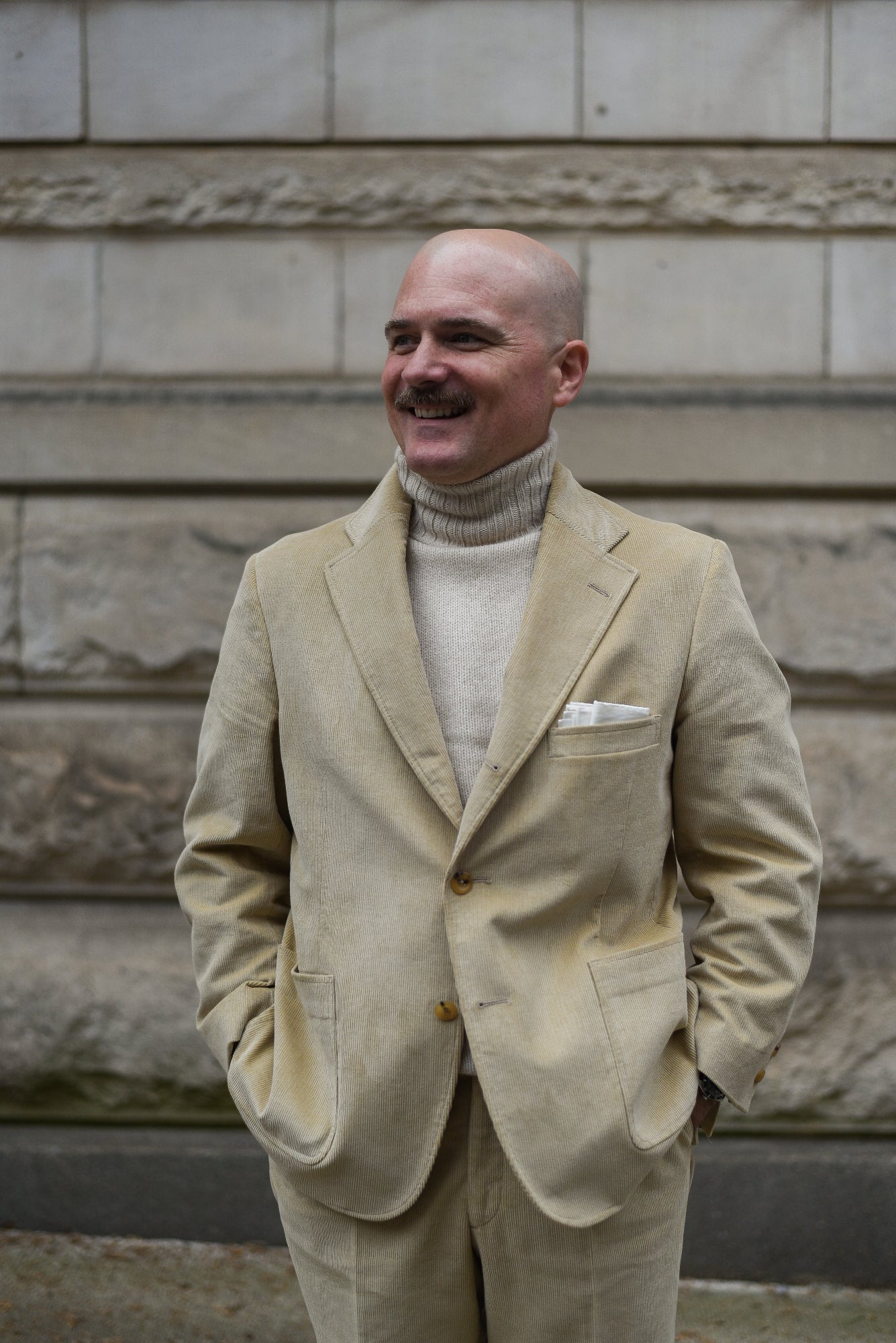 Man in a beige suit standing against a stone wall.