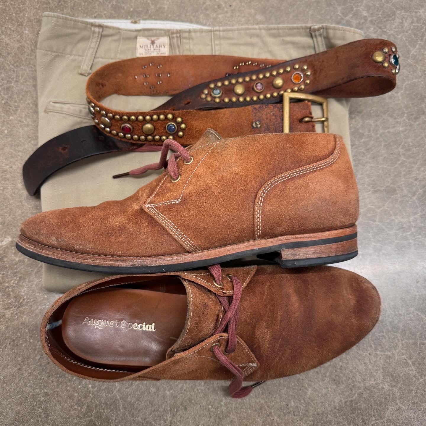 A pair of brown suede chukka-style boots with burgundy laces, displayed on top of a tan pair of khaki pants and a brown studded leather belt. The insole of the lower boot shows the brand name 'August Social'.