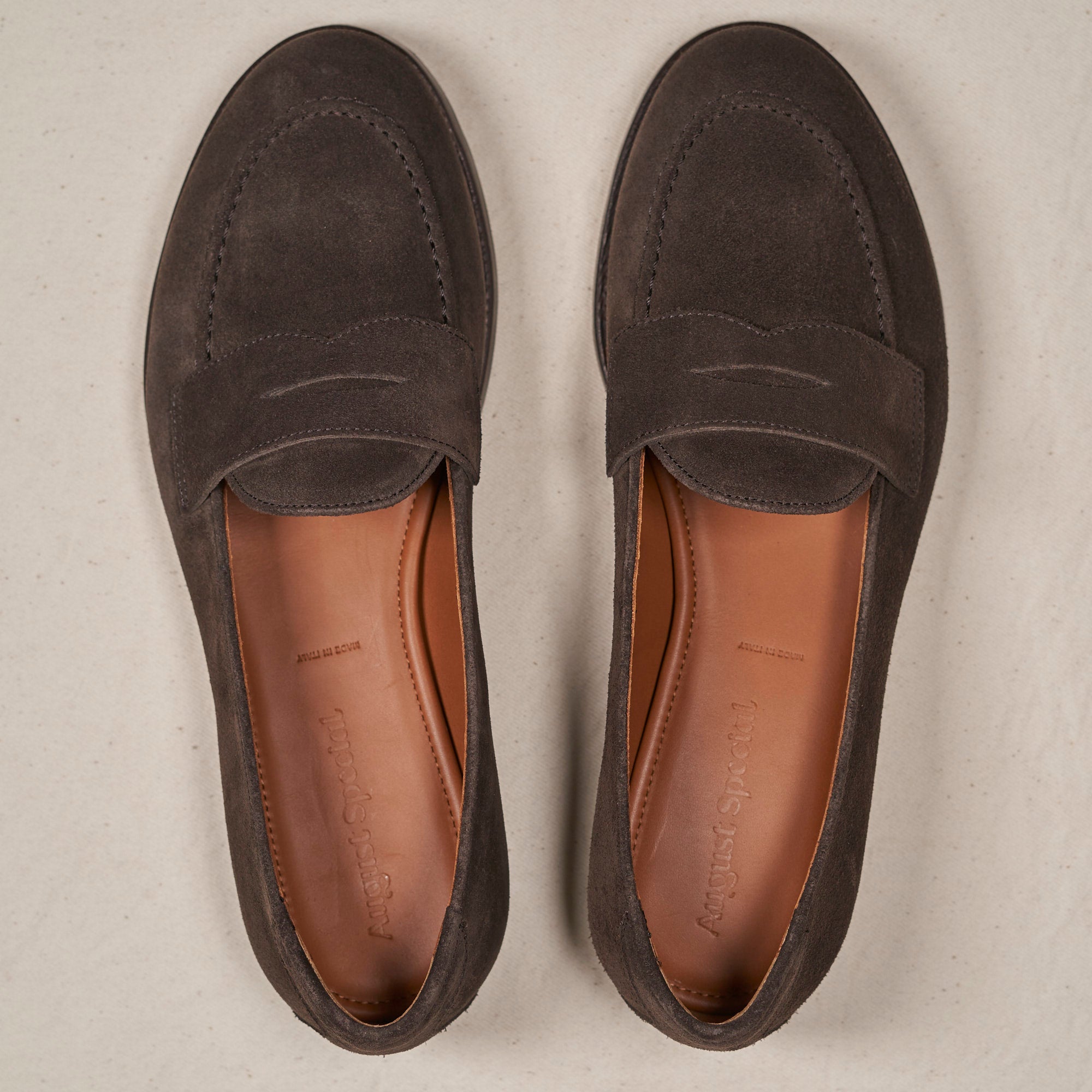 A pair of dark gray suede penny loafers shown from a top-down view on a light beige textured background.