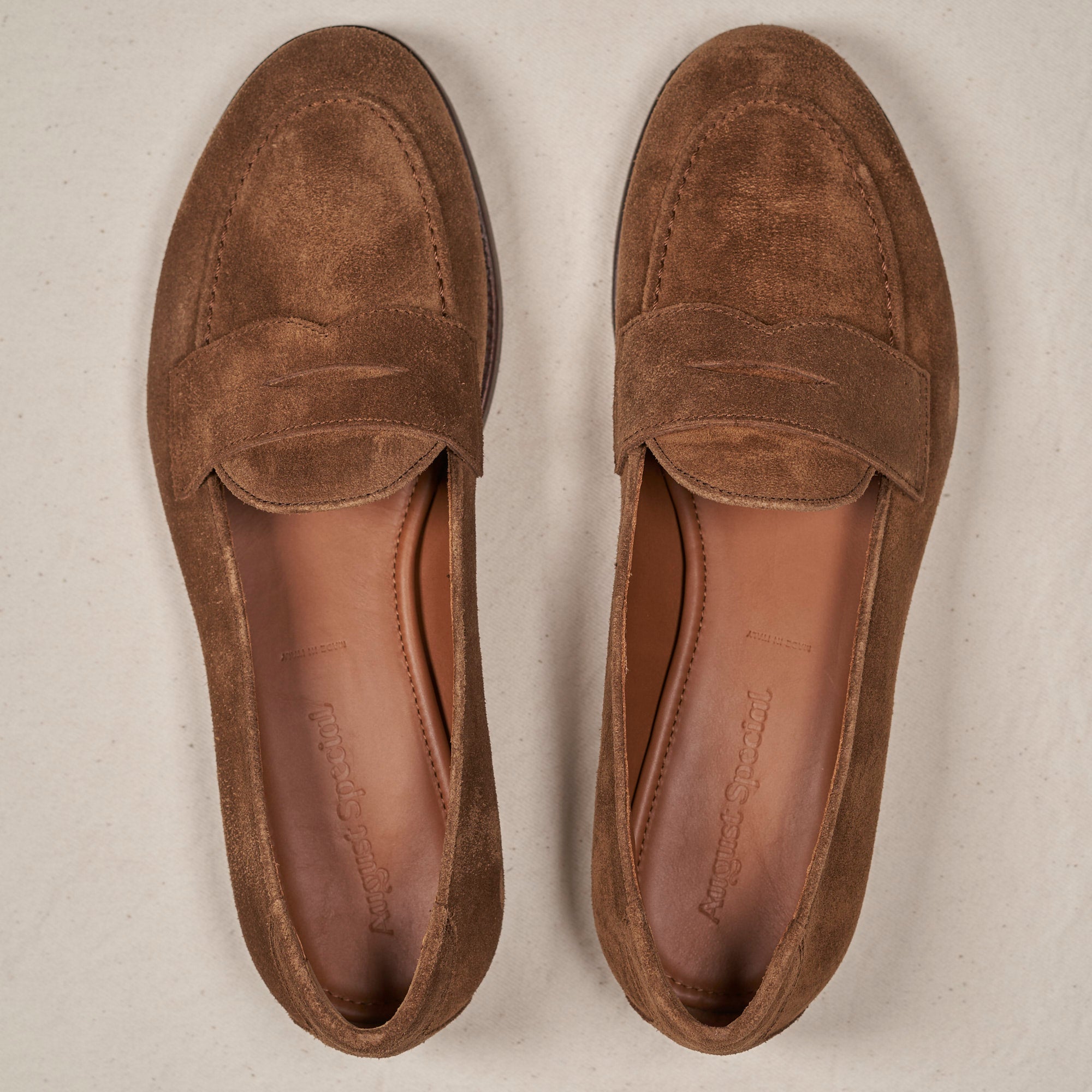A pair of brown suede penny loafers shown from a top-down view on a light beige textured background.
