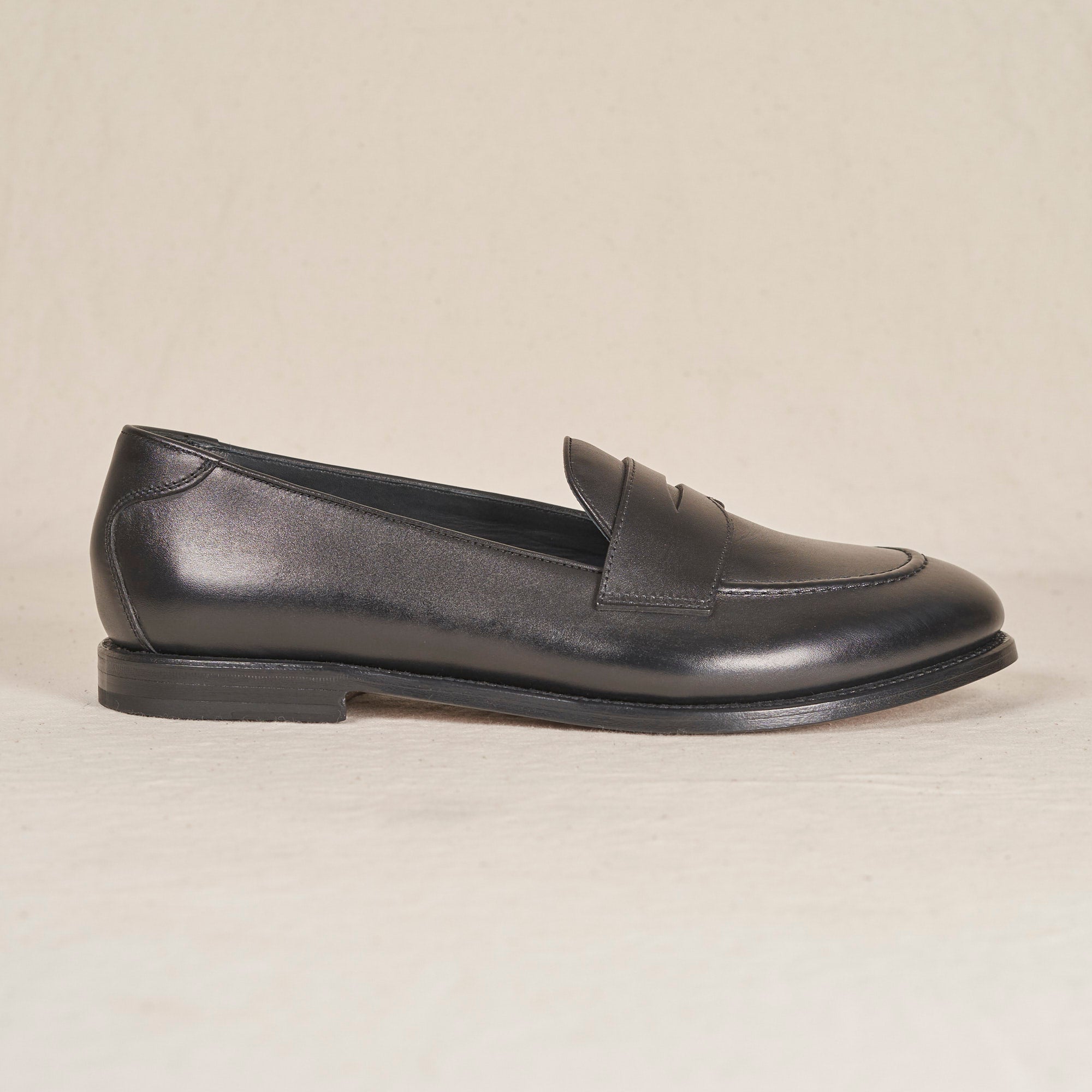 A single black leather penny loafer shoe shown in a side profile view against a light beige textured background.