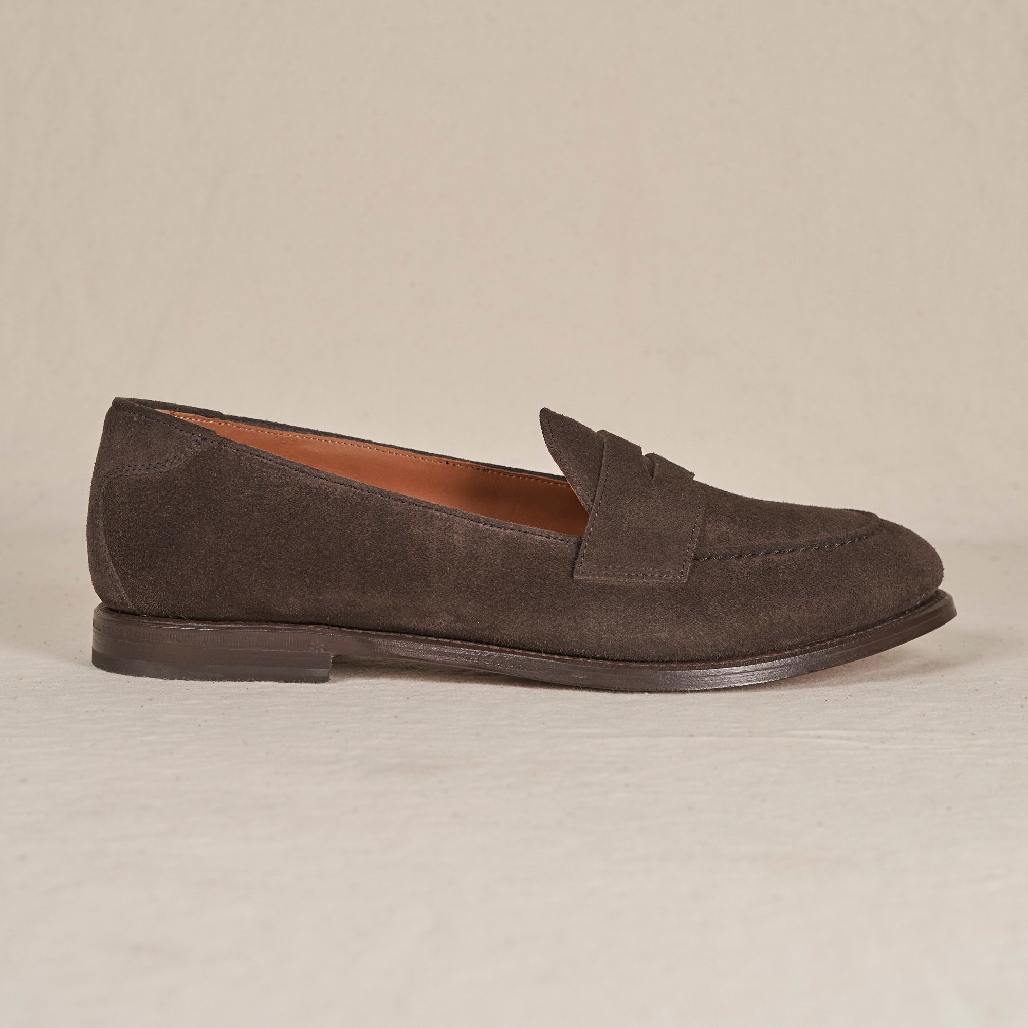 A single dark brown suede penny loafer shown in profile against a neutral, light beige textured background.