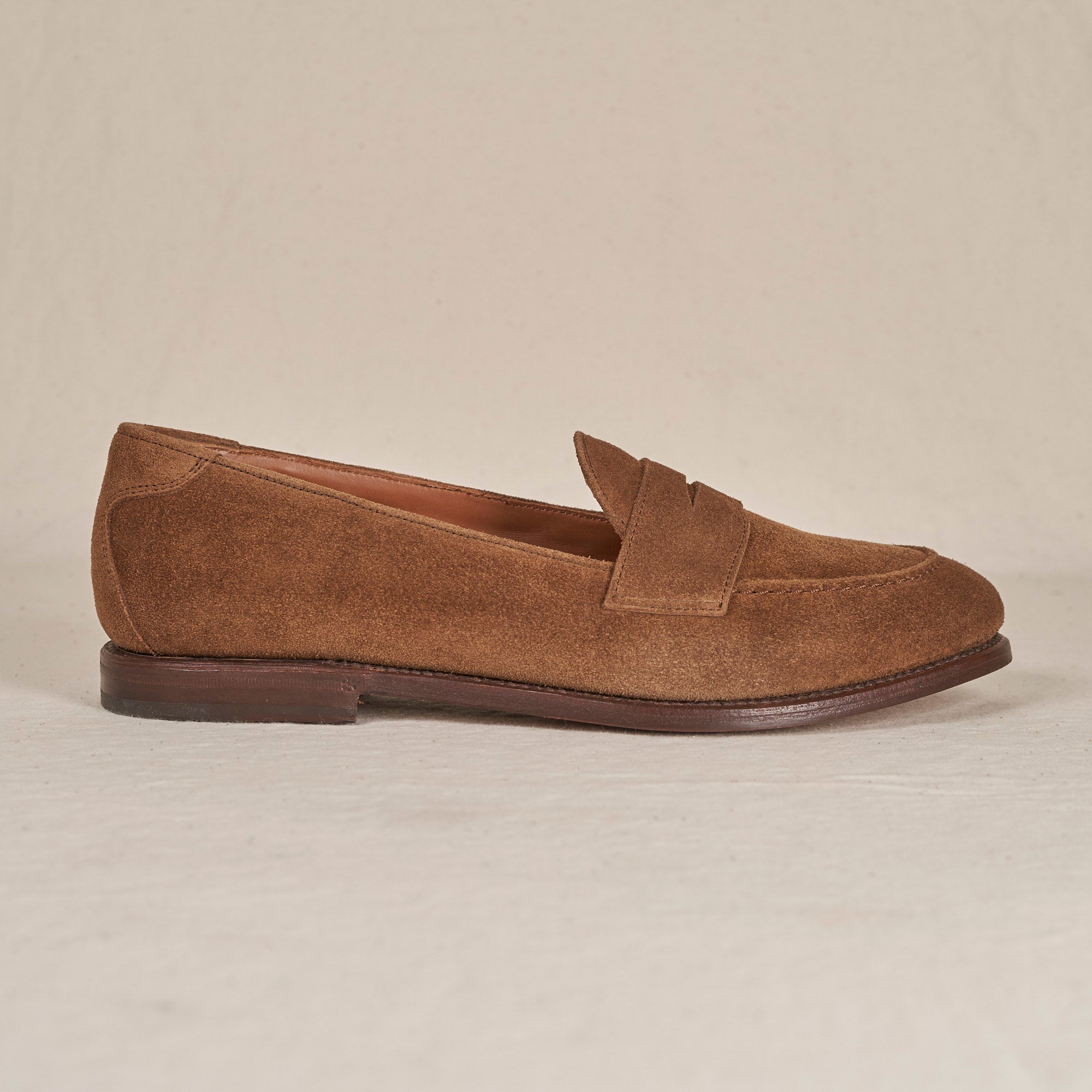A single brown suede penny loafer shown in profile against a neutral, light beige background.