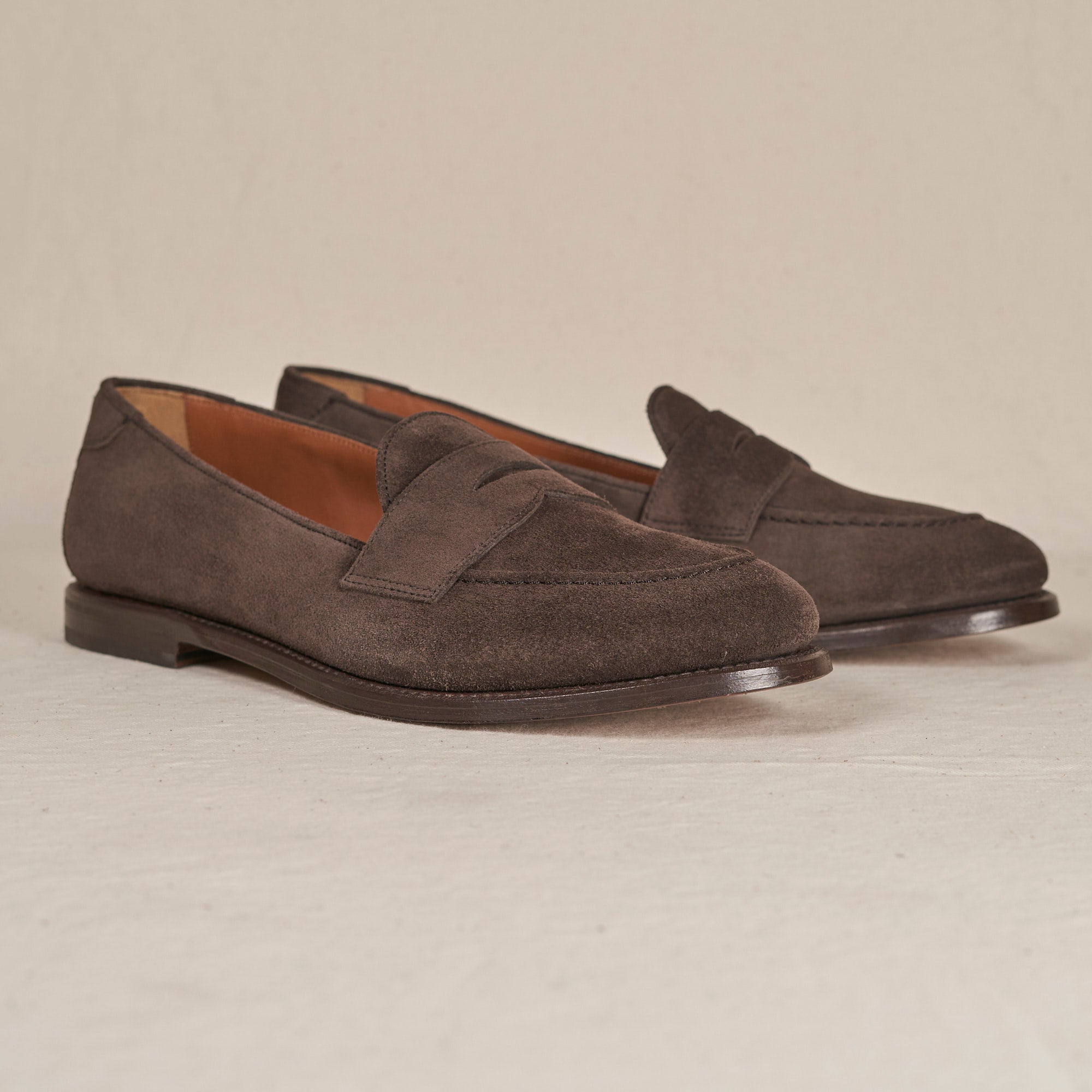 A pair of dark brown suede penny loafers with a tan interior lining, displayed on a light beige textured background.
