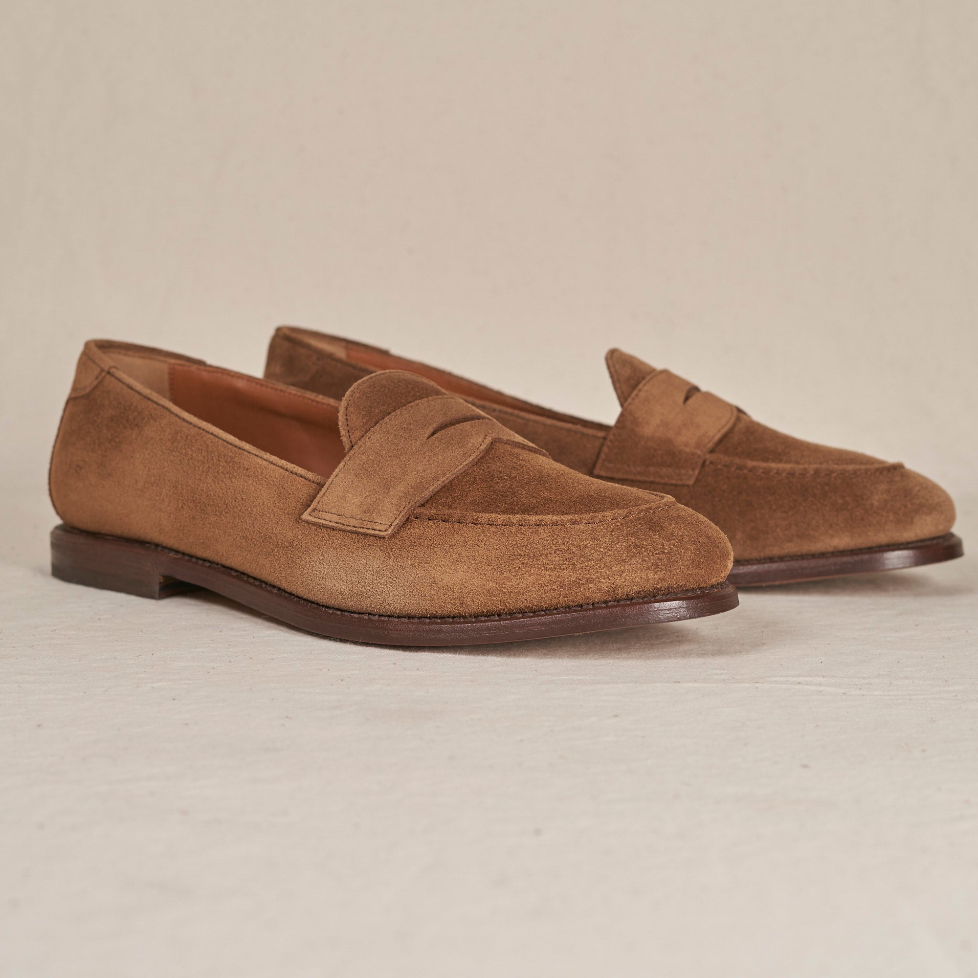 A pair of men's brown suede penny loafers with a classic strap across the vamp, displayed on a light beige textured studio background.
