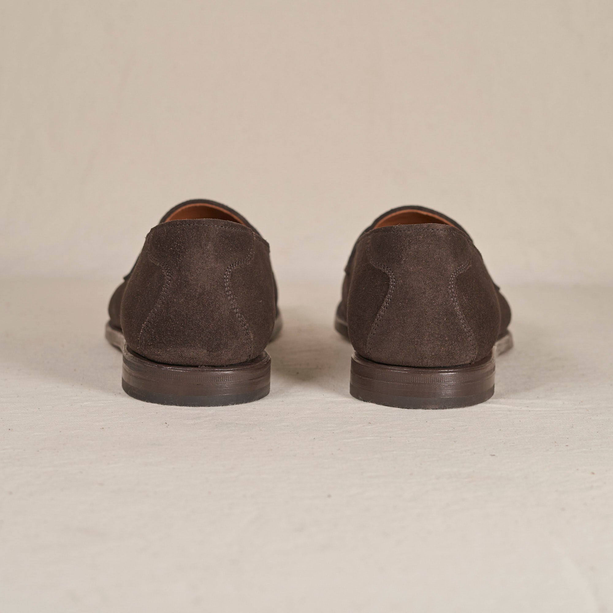 A pair of dark brown suede loafers shown from the back, positioned side-by-side on a light beige textured surface against a matching neutral background.