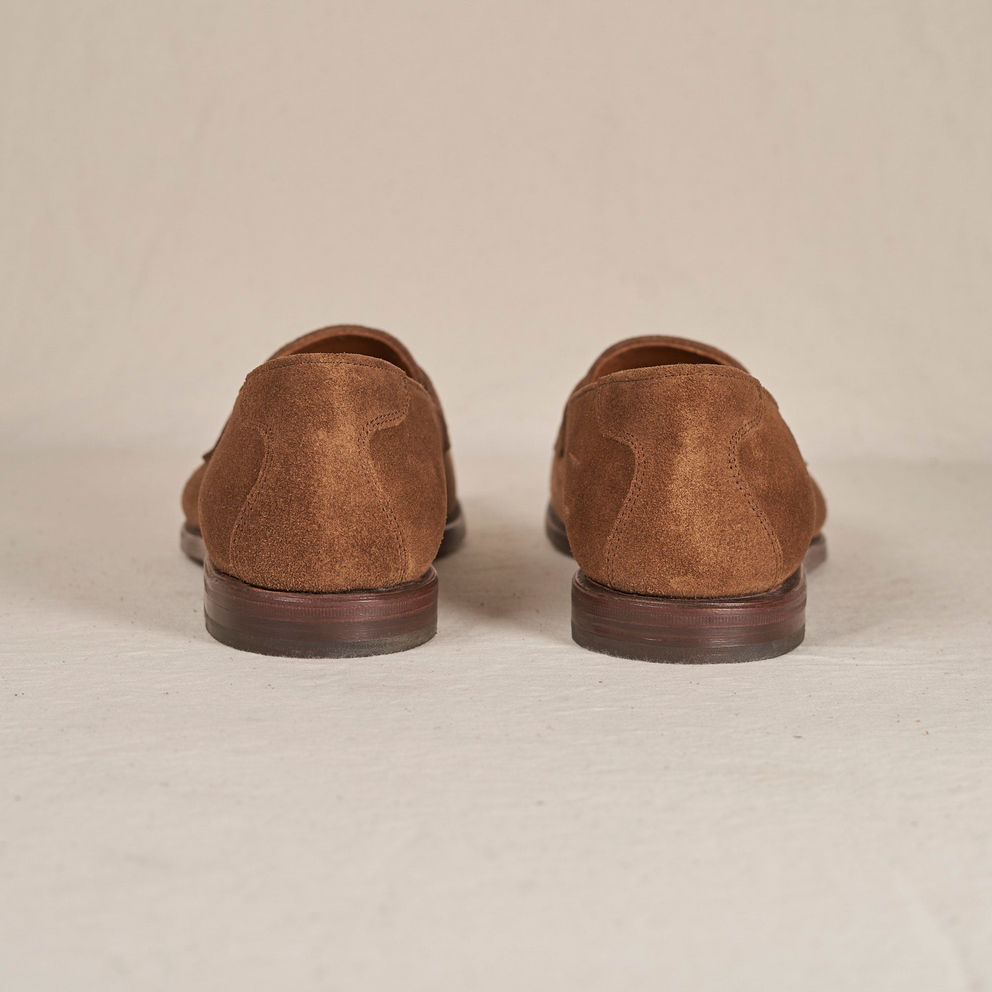 A pair of brown suede oxford shoes shown from the back, positioned side-by-side on a light beige textured surface.