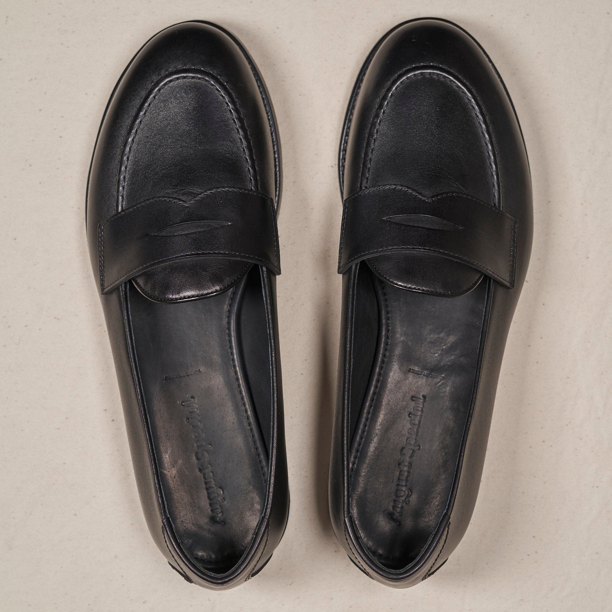 A pair of black leather penny loafers shown from a top-down view on a light beige textured background.