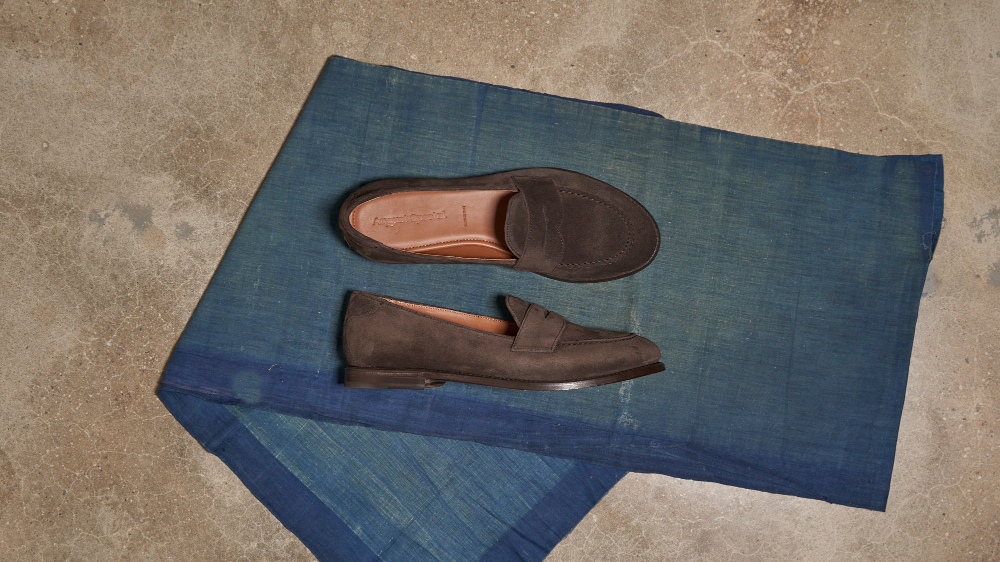 A pair of dark brown suede penny loafers displayed on a folded blue textile over a textured concrete floor.