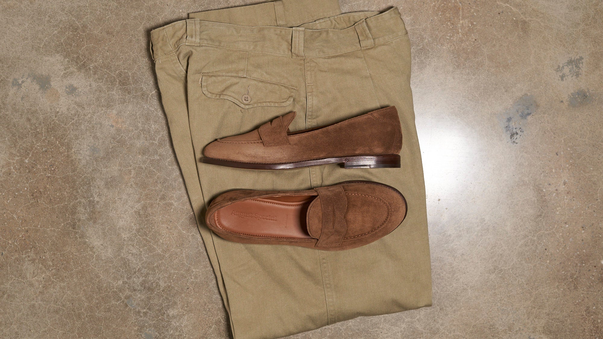 A pair of brown suede penny loafers arranged on top of folded khaki pants, photographed from above on a textured concrete floor.