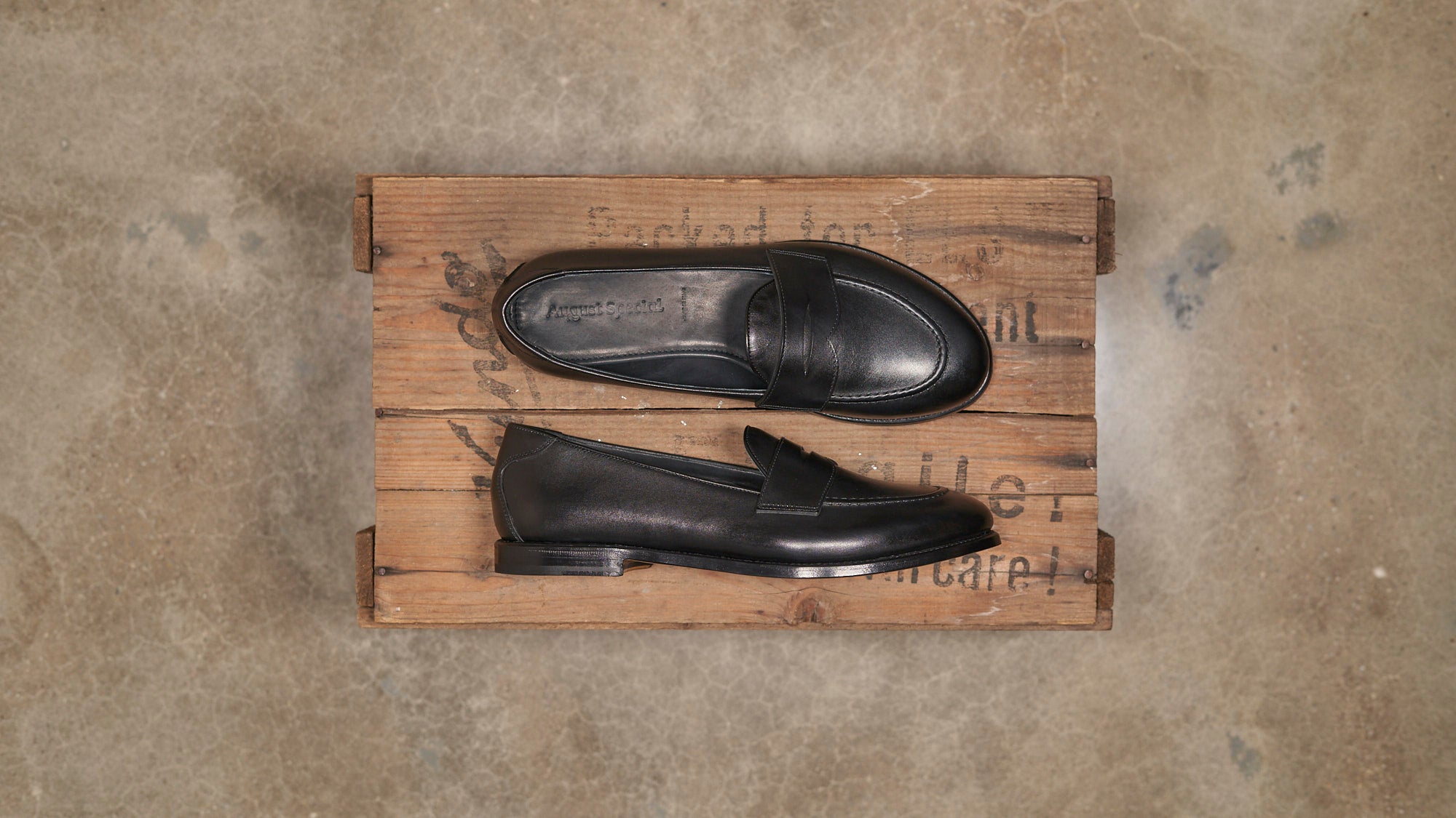 A pair of black leather penny loafers displayed on a rustic wooden crate against a textured concrete floor.
