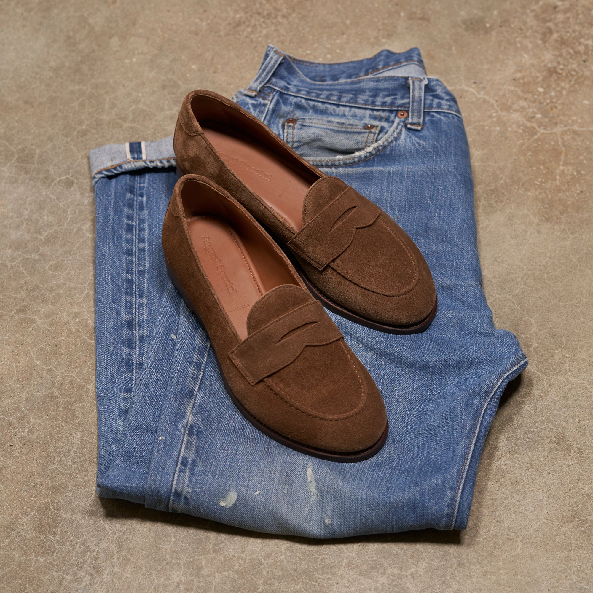 A pair of brown suede penny loafers placed on top of folded blue jeans on a textured concrete floor.
