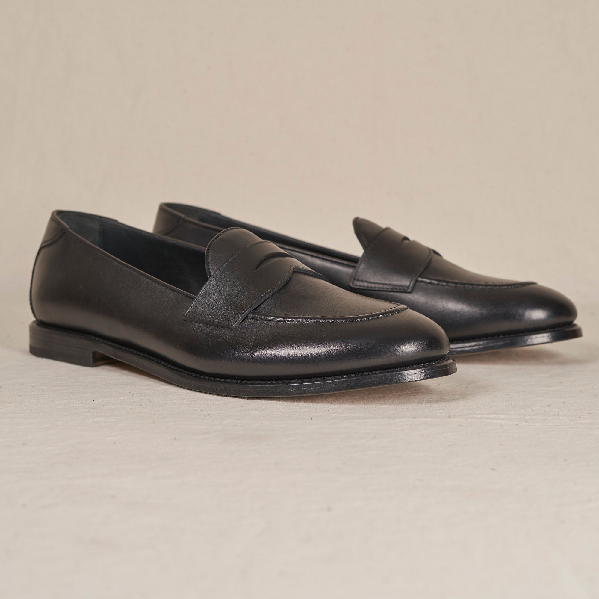 A pair of black leather penny loafers with a smooth finish, displayed on a light beige textured surface against a matching neutral background.