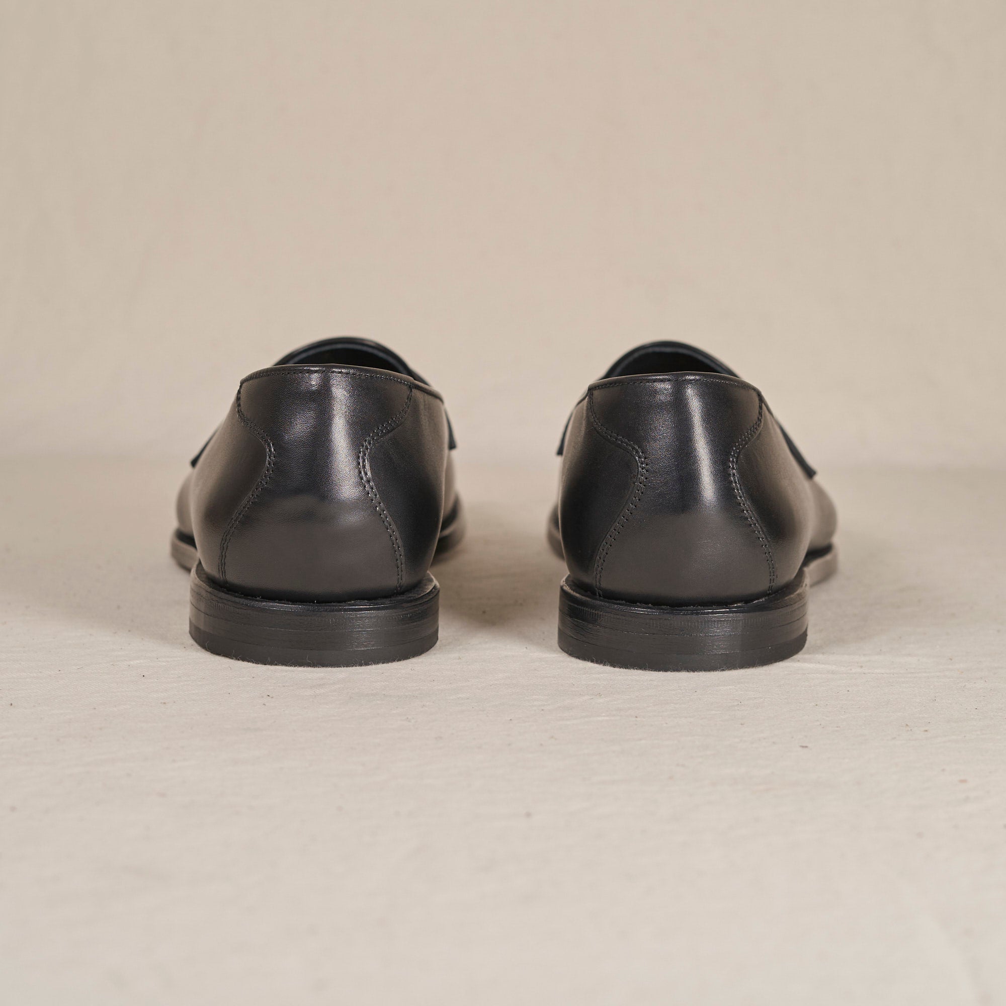 A pair of black leather dress shoes shown from the back, positioned side-by-side on a light beige textured surface.