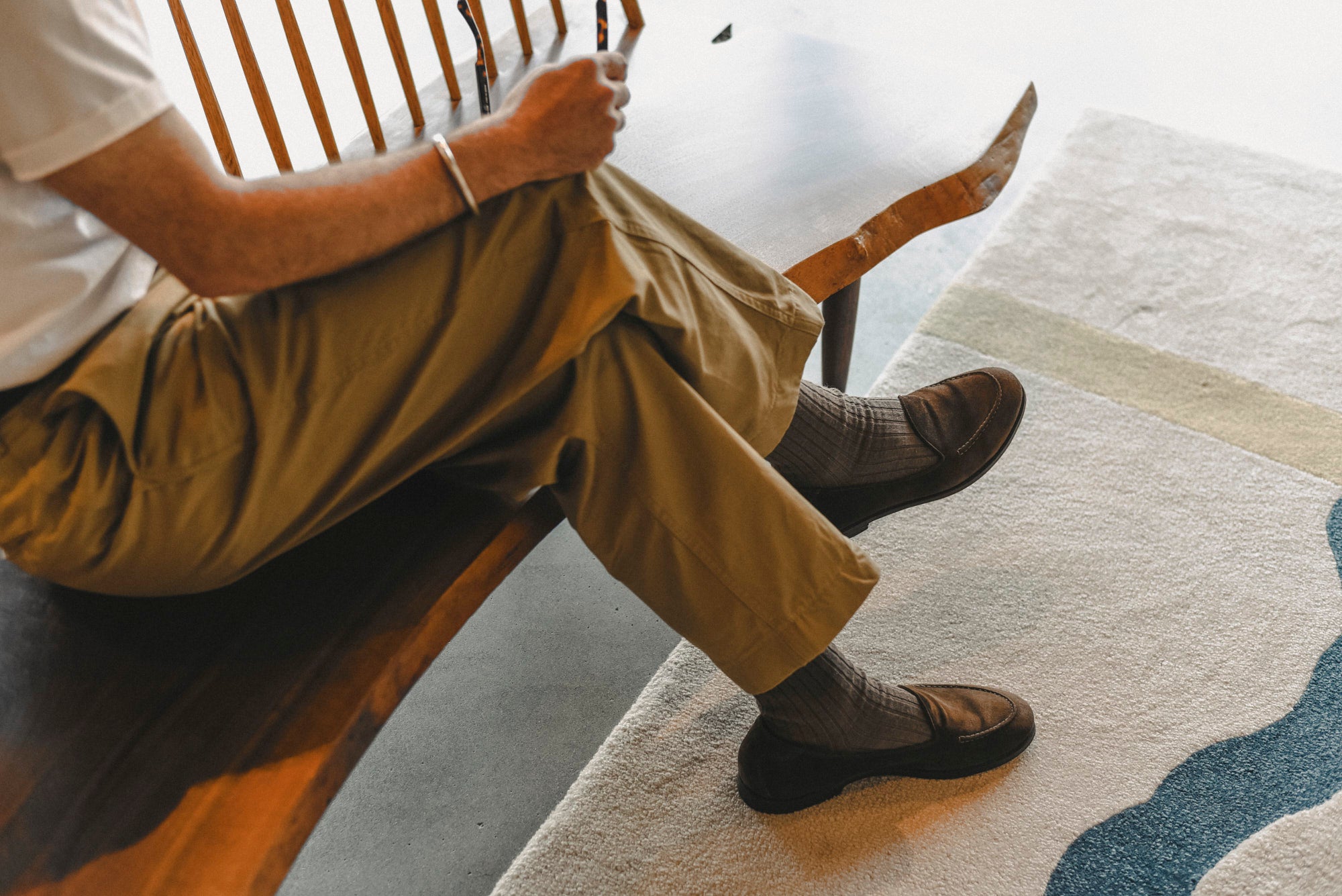 A person seated on a wooden bench indoors, wearing khaki pants, dark socks, and dark slip-on shoes, with a patterned rug visible on the floor.