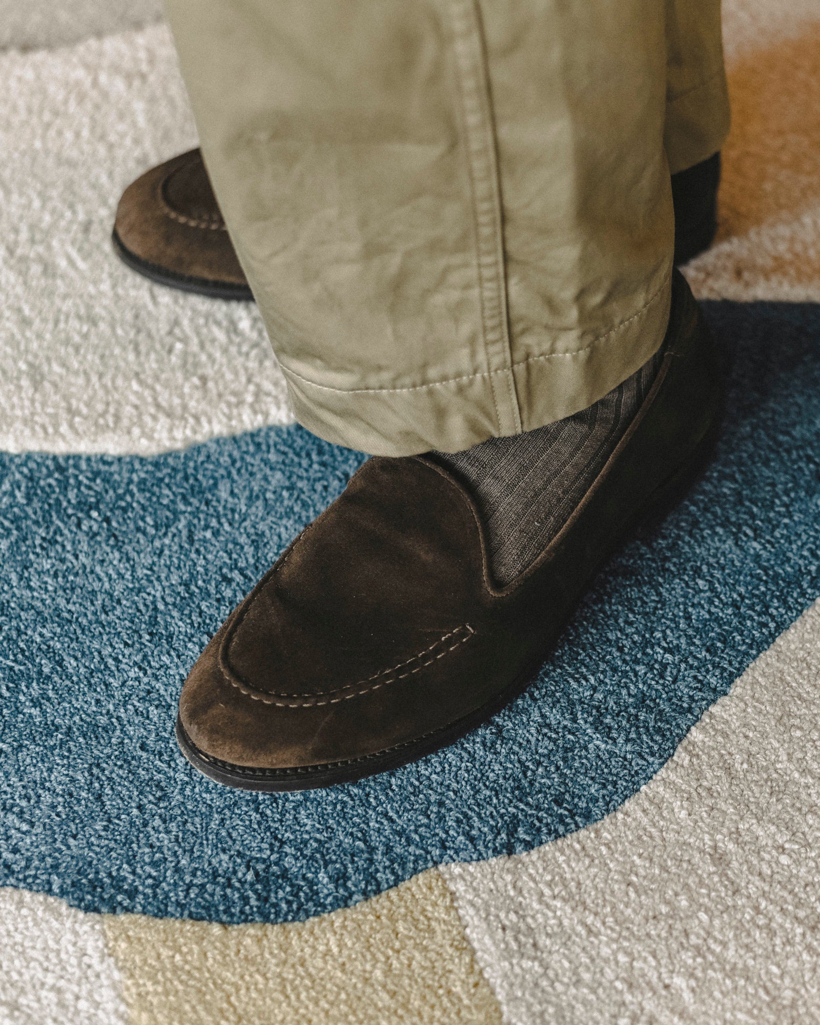 A close-up of a person wearing dark brown suede slip-on loafers with olive green pants and a blue and beige patterned rug.