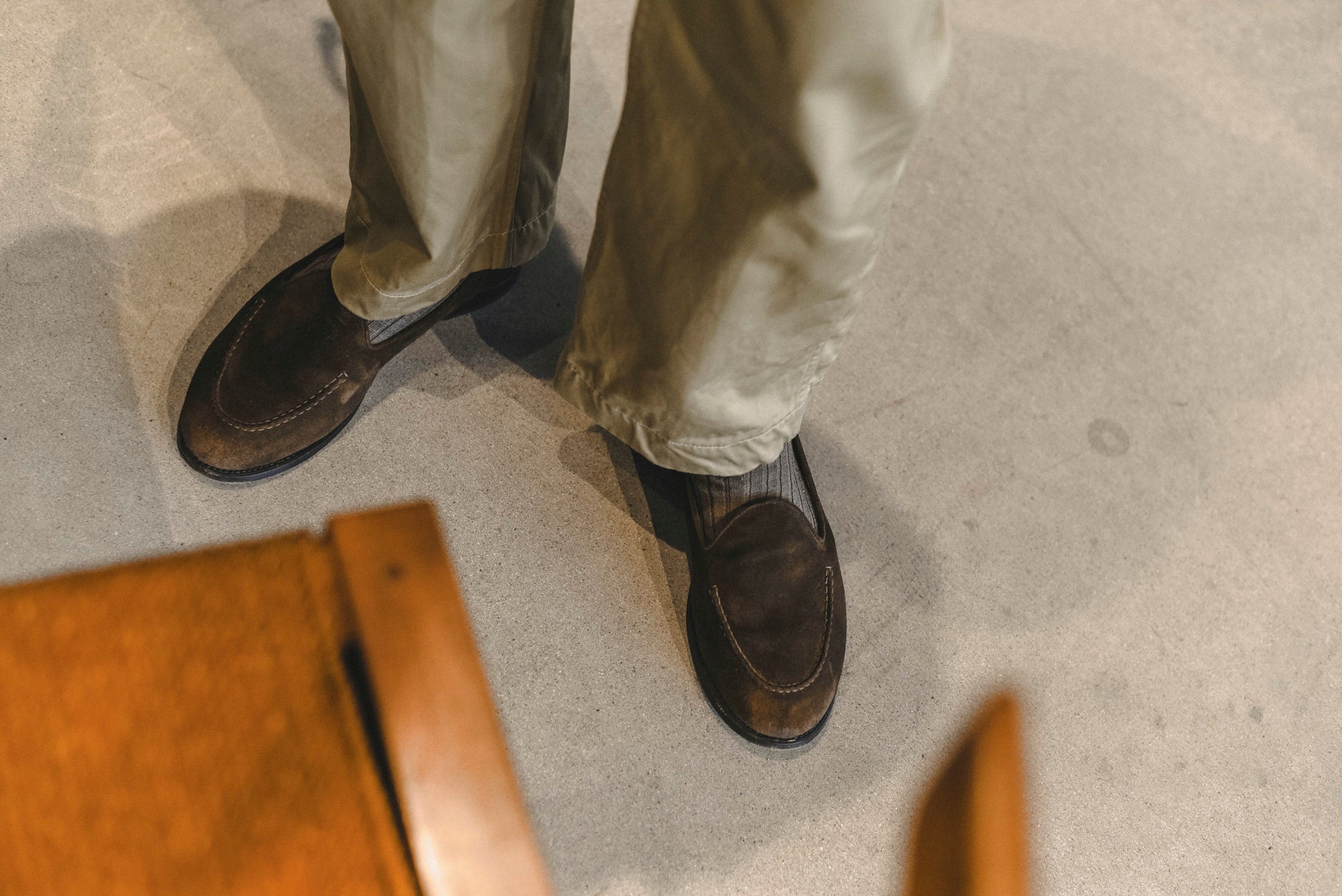 A close-up, high-angle view of a person's legs and feet wearing dark brown suede loafers and light beige trousers, standing on a concrete floor.