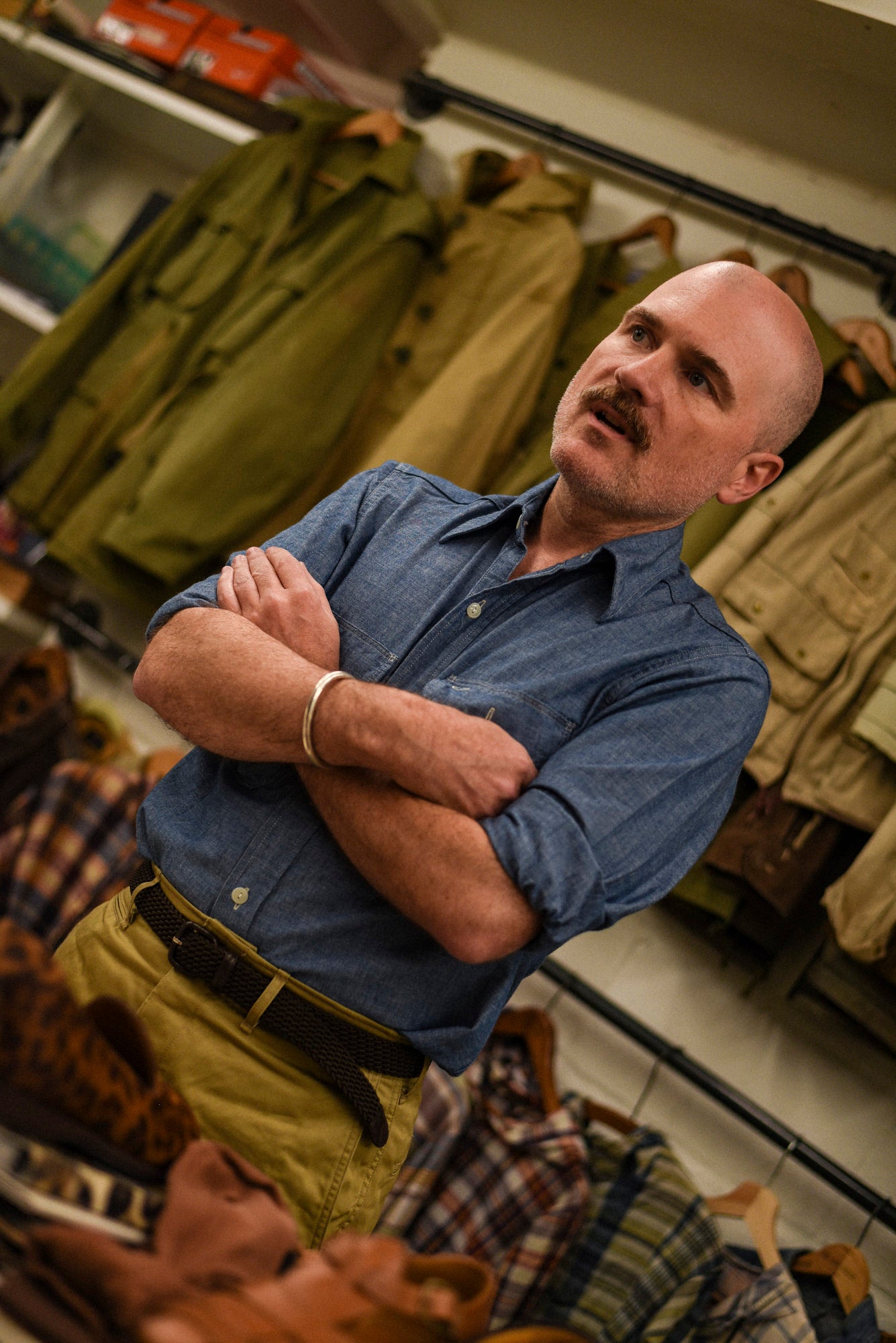 A man with a mustache wearing a blue button-down shirt and khaki pants, standing with arms crossed in a clothing store with jackets hanging in the background.