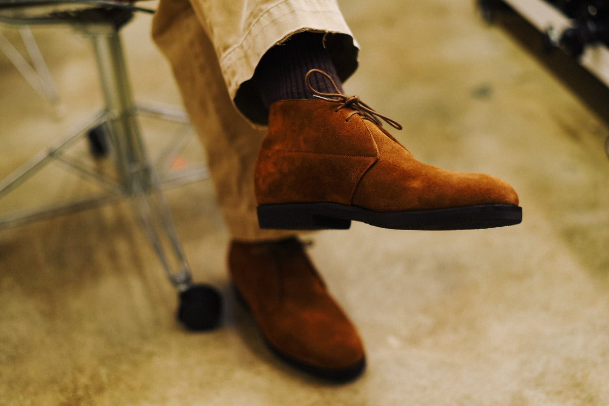 A person wearing brown suede chukka boots with dark socks and khaki pants, shown in a close-up lifestyle shot on a concrete floor.