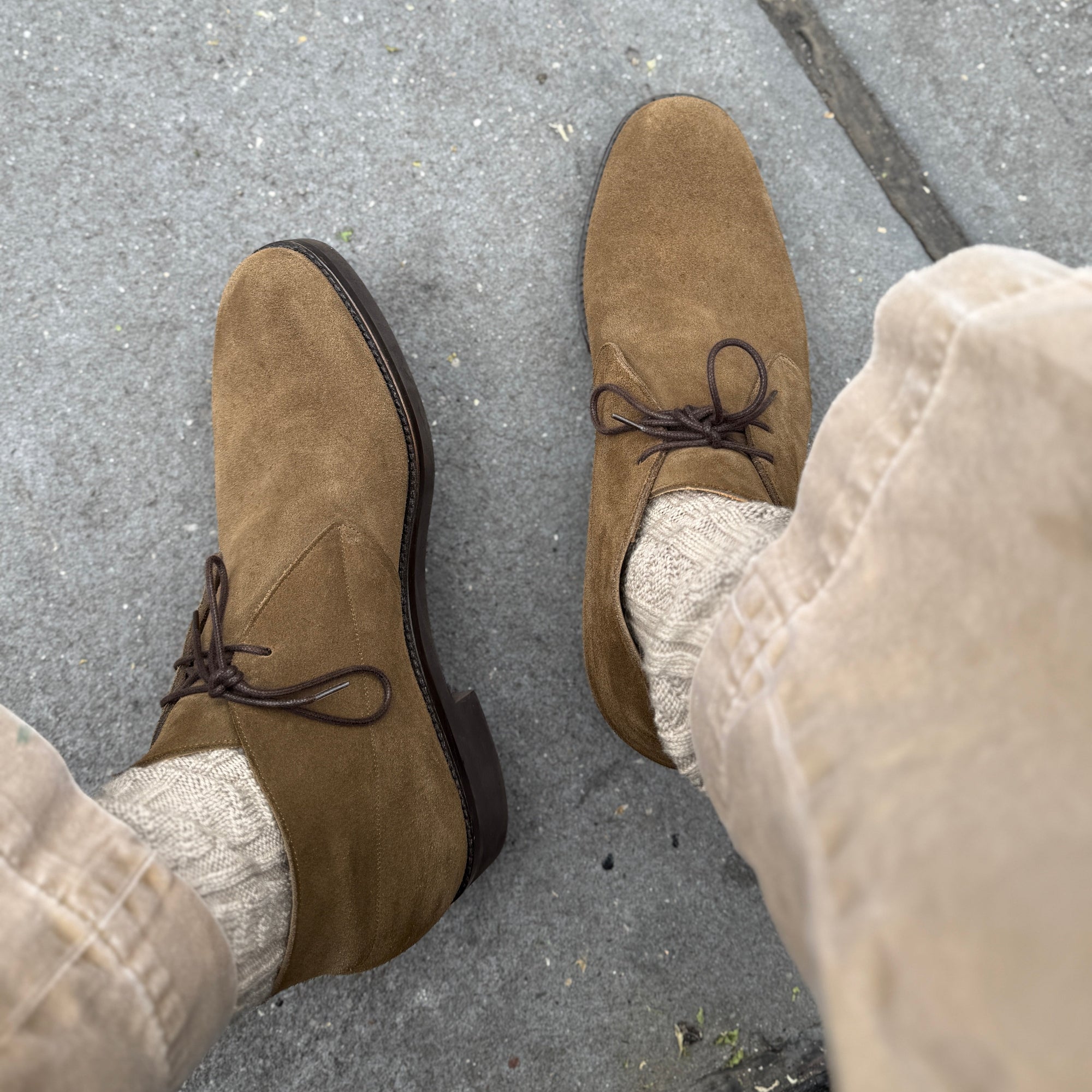 A top-down view of a person wearing brown suede derby-style shoes with dark laces, paired with cream socks and beige pants, standing on a gray concrete pavement.