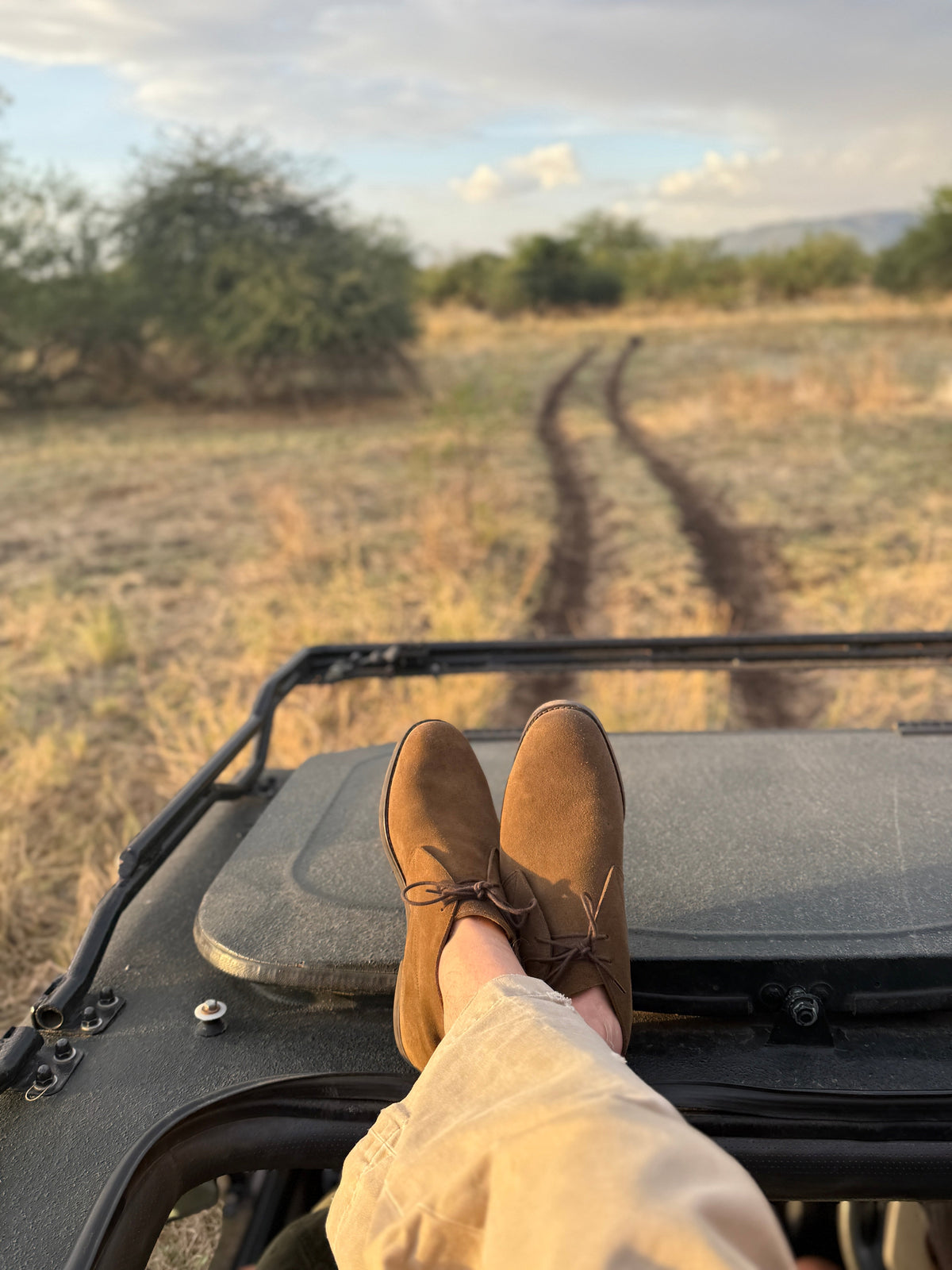 A person's feet in tan suede lace-up boots and beige pants resting on the back of a safari vehicle, looking out over a dry grassy landscape with a dirt track leading into the distance.